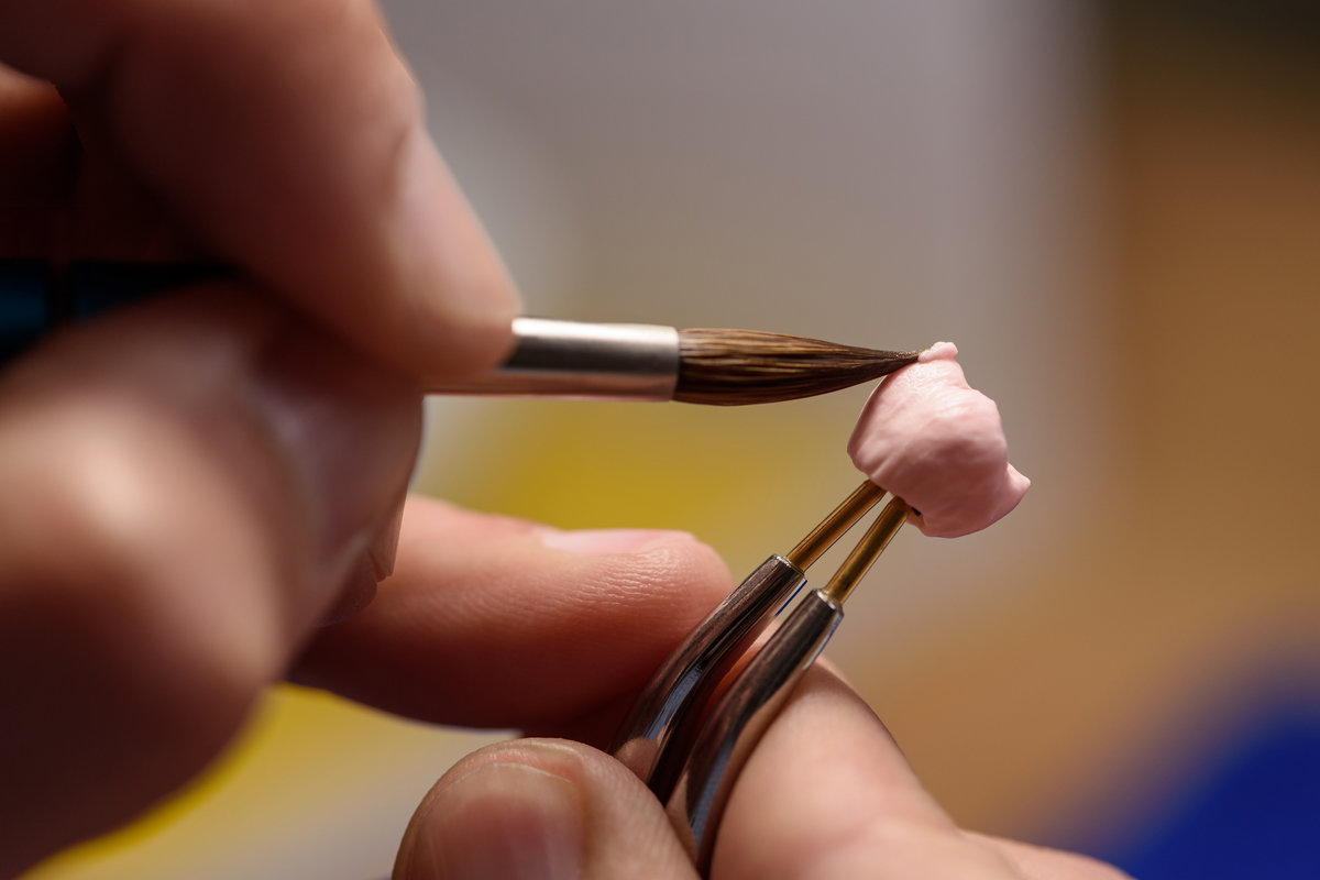 Dental prosthesis work. Teeth painting. Dental technician working with tooth denture at prosthesis laboratory. Technician applies ceramic material to the crown of a dental implant using a brush.